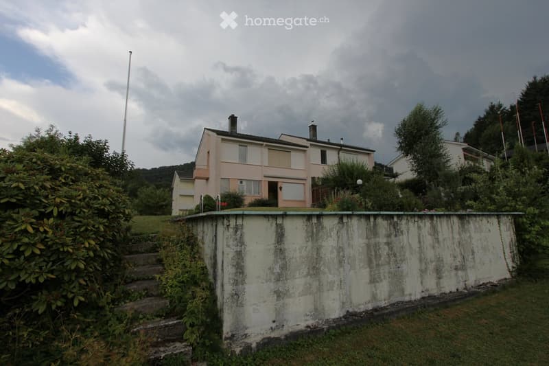 Two-story house with beige and white exterior, multiple windows, and a chimney, surrounded by greenery and a raised ground floor.