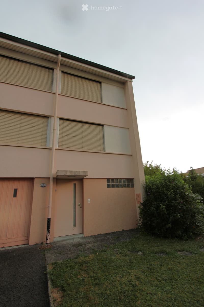 Two-story house with beige exterior, multiple windows with blinds, garage door, and small garden area.