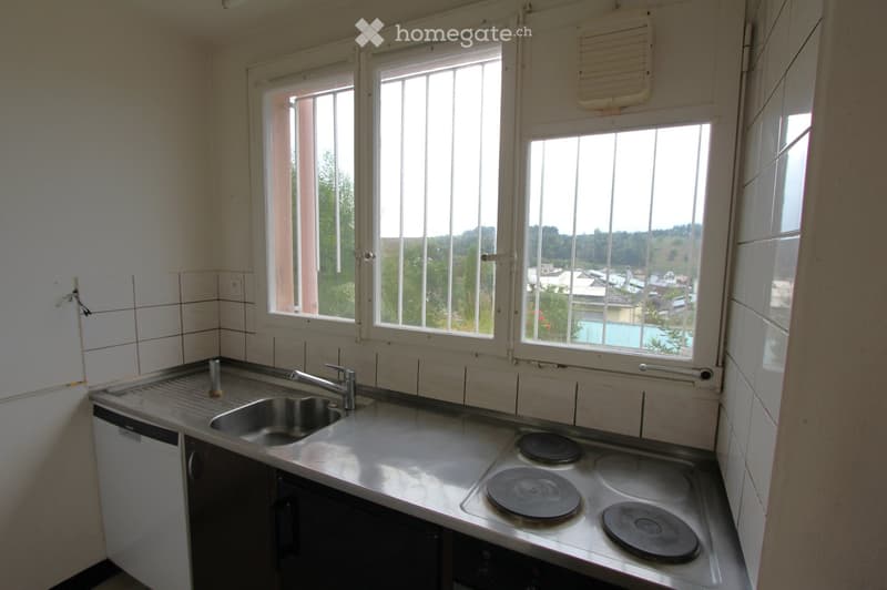 Empty kitchen with stainless steel sink, faucet, electric stove, and dishwasher, large windows overlooking a green area.