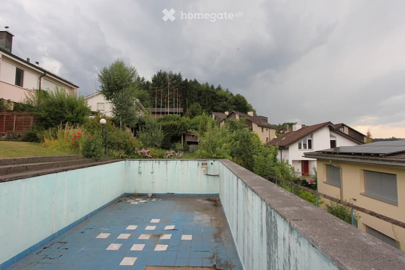 An abandoned rooftop area with a blue tiled floor and light blue walls, surrounded by residential houses and greenery.