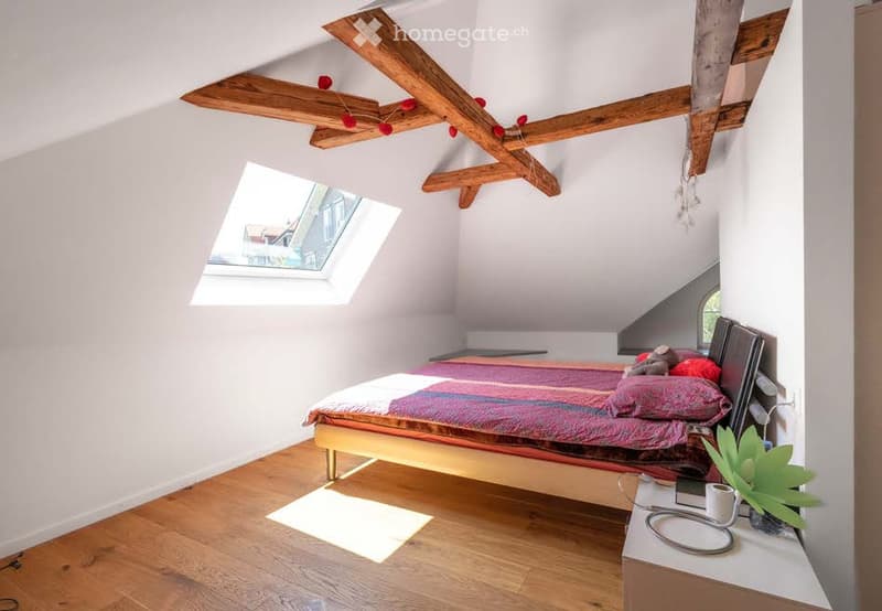Cozy attic bedroom with wooden beams, skylight window, bed with colorful bedding, nightstand with plant, and mirror on wall.