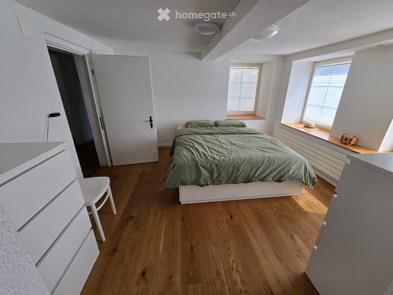 Cozy bedroom with wooden flooring, a bed with green bedding, a white chair, and a small window seat with a hat and suitcase.
