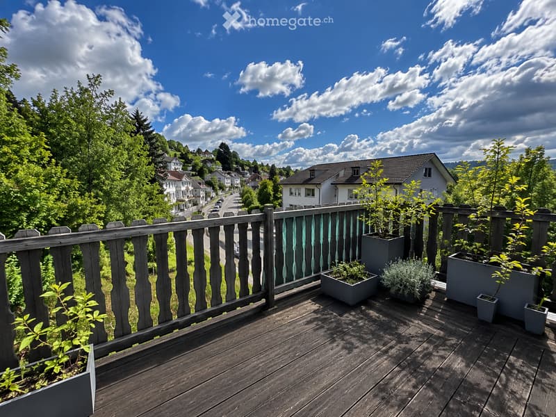 Wooden balcony with railing, potted plants, overlooking a scenic town with houses and greenery under a partly cloudy sky.