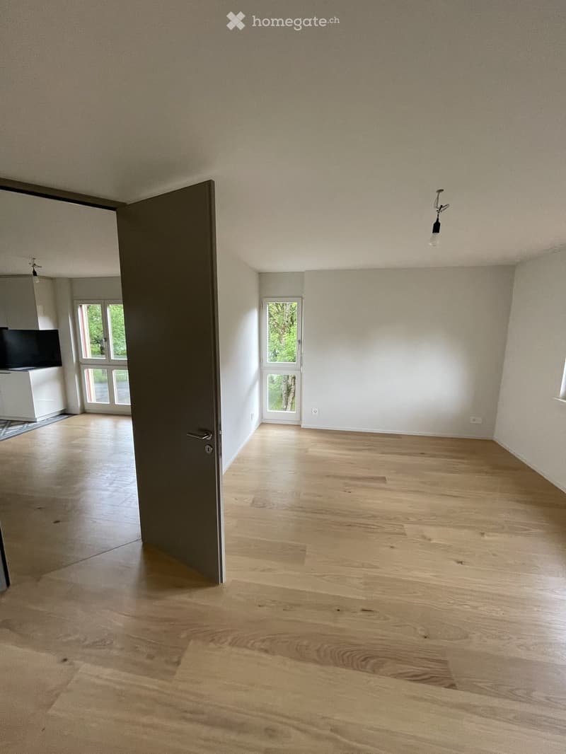 Empty room with light wooden flooring, white walls, and a closed door leading to another space. Natural light streams in from a window in the adjacent room.