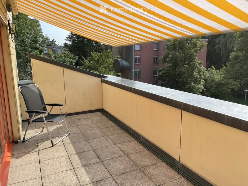 Balcony with tiled floor, yellow walls, a black folding chair, and a striped awning providing shade.