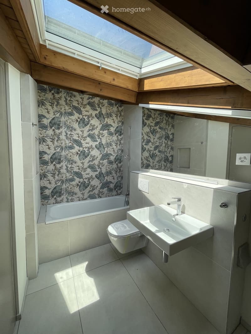 Modern bathroom with a bathtub, toilet, sink, and floral shower curtain. Features skylight windows and wooden beams on the ceiling.