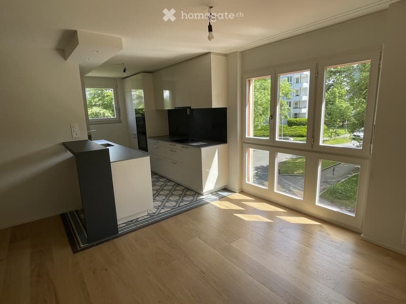 Modern kitchen with light-colored cabinets, a central island, and large windows allowing natural light to fill the space. The floor is wooden, and the countertops are sleek and contemporary.