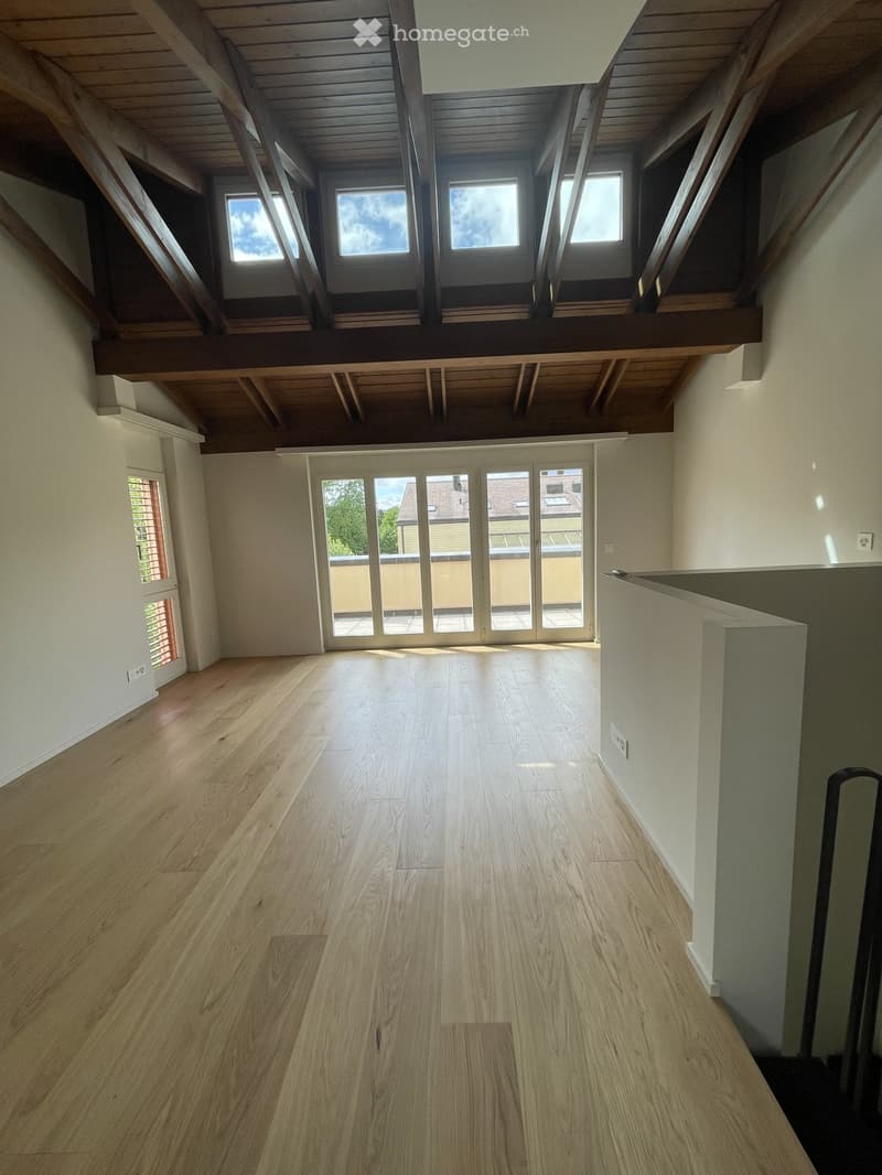Empty room with wooden flooring, white walls, large windows, and a high, slanted wooden ceiling with exposed beams.