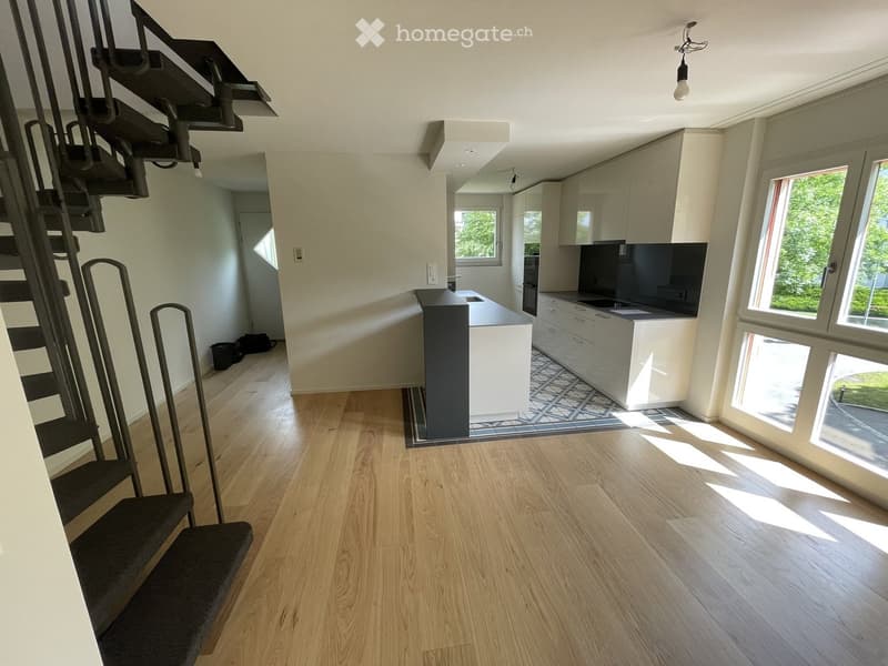 Open-plan living and kitchen area with light wooden flooring, modern kitchen island, cabinets, and a staircase leading to another level.