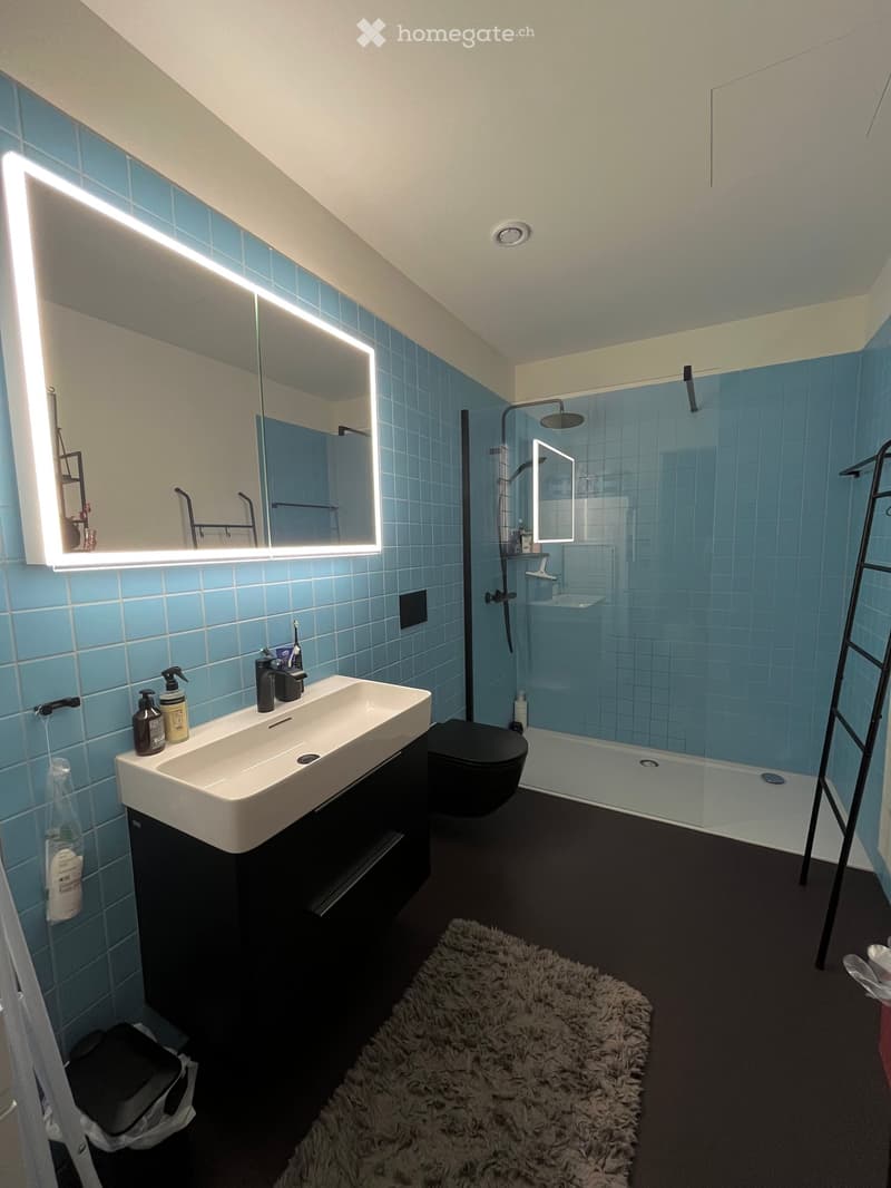 Modern bathroom with blue tiles, a large illuminated mirror, a white sink with a black cabinet, a toilet, a shower area with a glass enclosure, and a fluffy white rug on the floor.