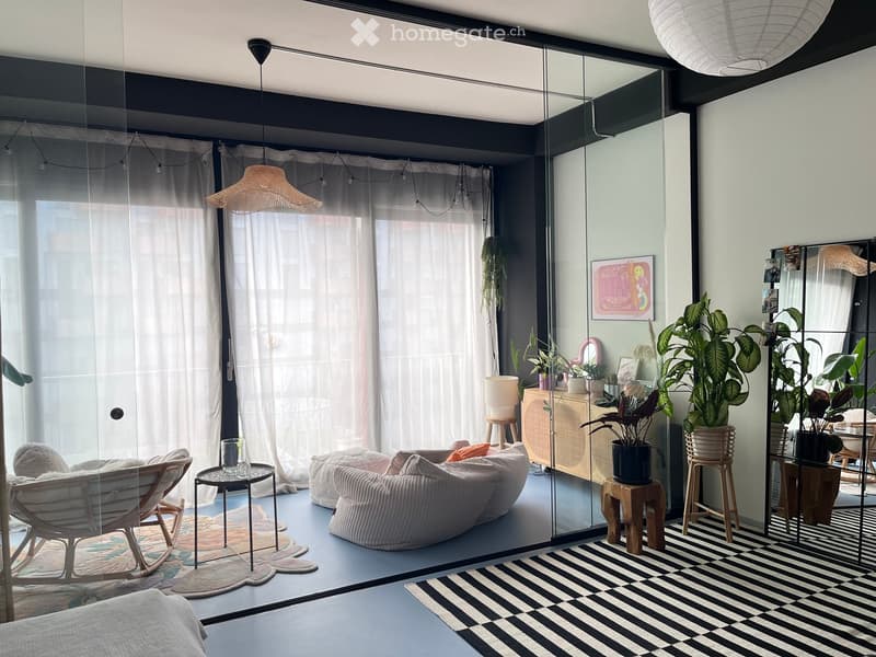 Modern living room with a sofa, chair, small table, and various plants. Features a large mirror, hanging lamps, and a mix of patterned and striped rugs.