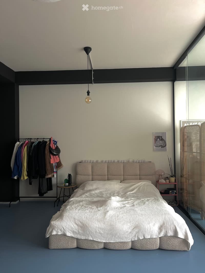 Modern bedroom with a large bed, upholstered headboard, nightstand, and a clothing rack with hanging clothes. The room features a pendant light and a picture frame on the wall.