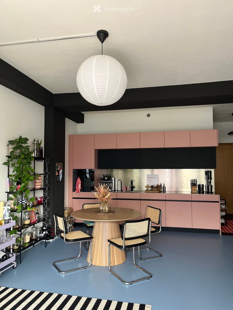 Modern kitchen with pink and black cabinets, a round wooden dining table with four chairs, a hanging spherical light fixture, and a potted plant on a shelf.