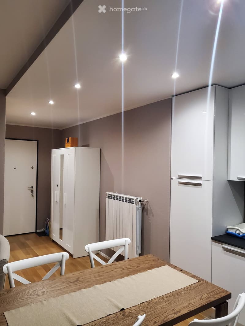 A modern, well-lit room with a wooden dining table and white chairs, a refrigerator, a cabinet, and a radiator. The room features a wooden floor and recessed ceiling lights.