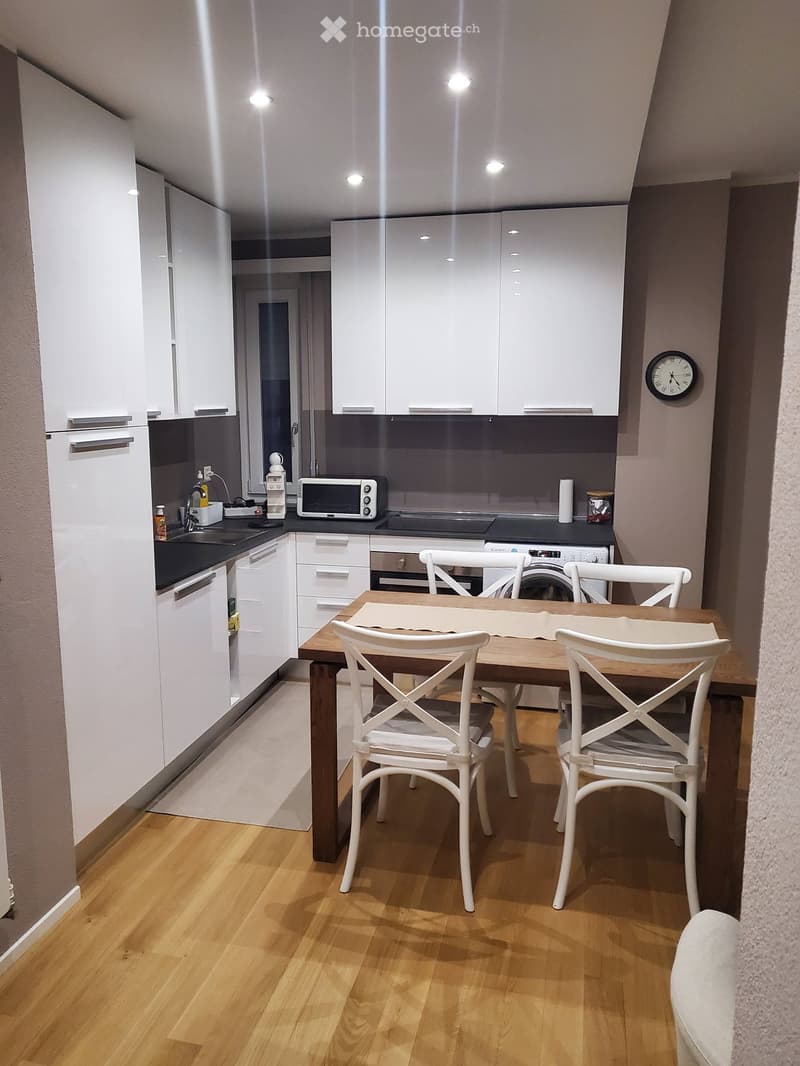 Modern kitchen with white cabinets, wooden floor, dining table with four white chairs, microwave, oven, fridge, sink, and faucet.
