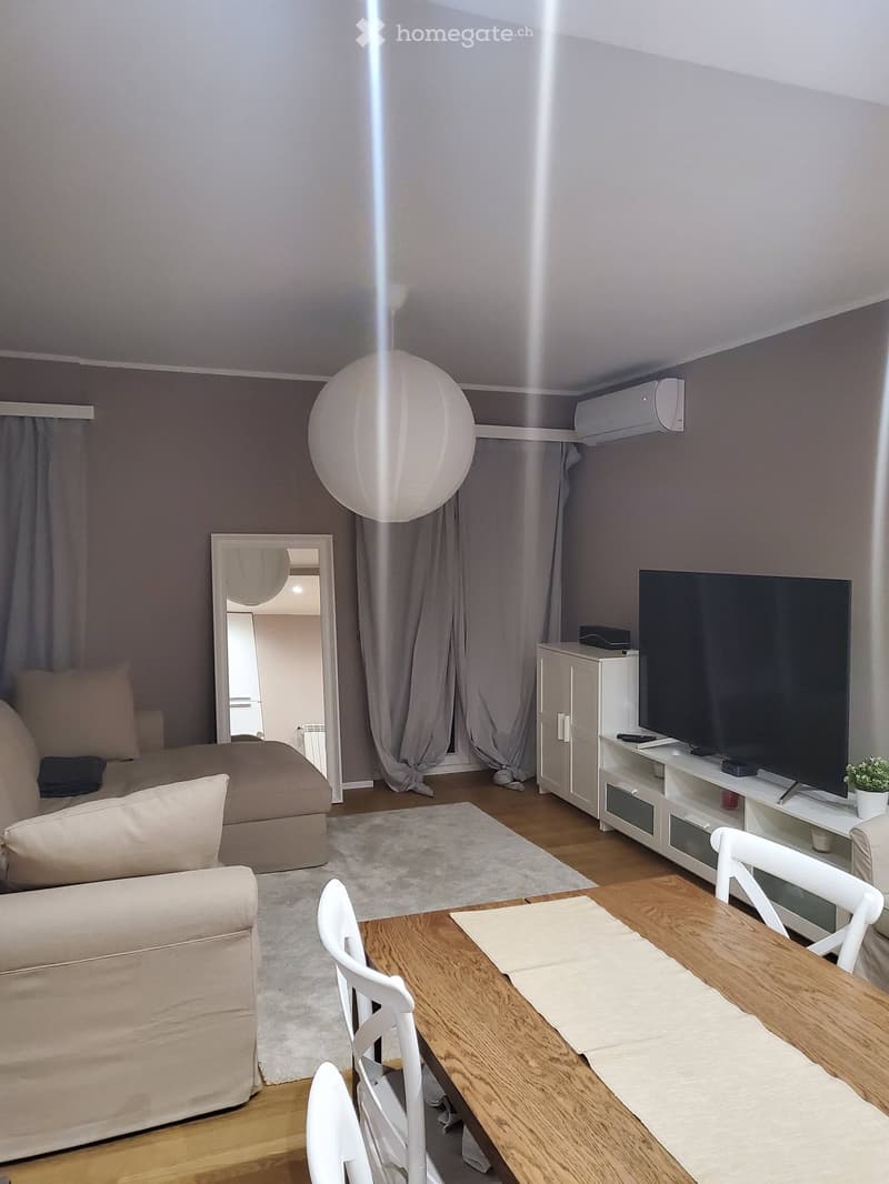 A modern living room with a sofa, a wooden dining table, white chairs, a television on a cabinet, a large round ball lamp, and a mirror leading to another room.