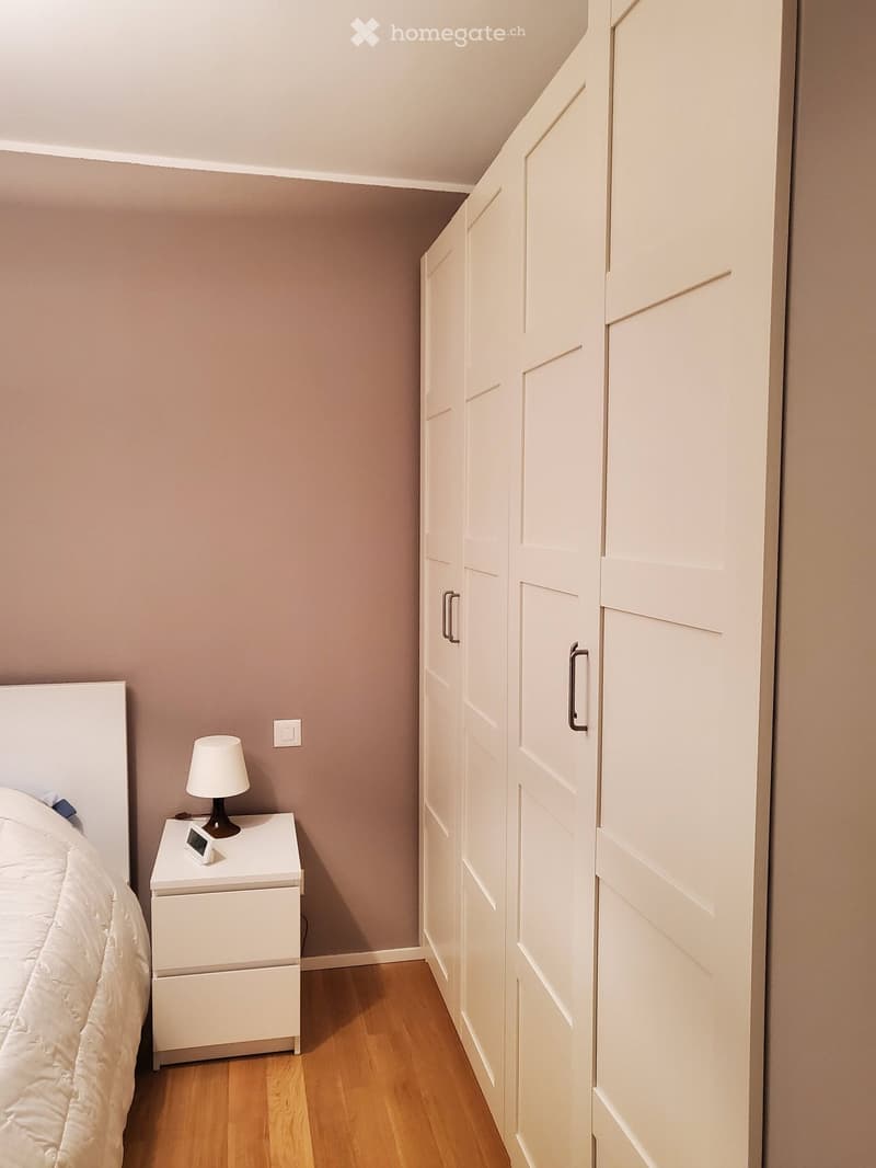 Modern bedroom with a white bed, nightstand, lamp, and large white wardrobe with mirrored doors.