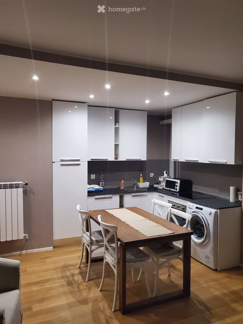 Modern kitchen with white cabinets, wooden floor, central dining table with four chairs, dishwasher, microwave, sink, and washing machine.