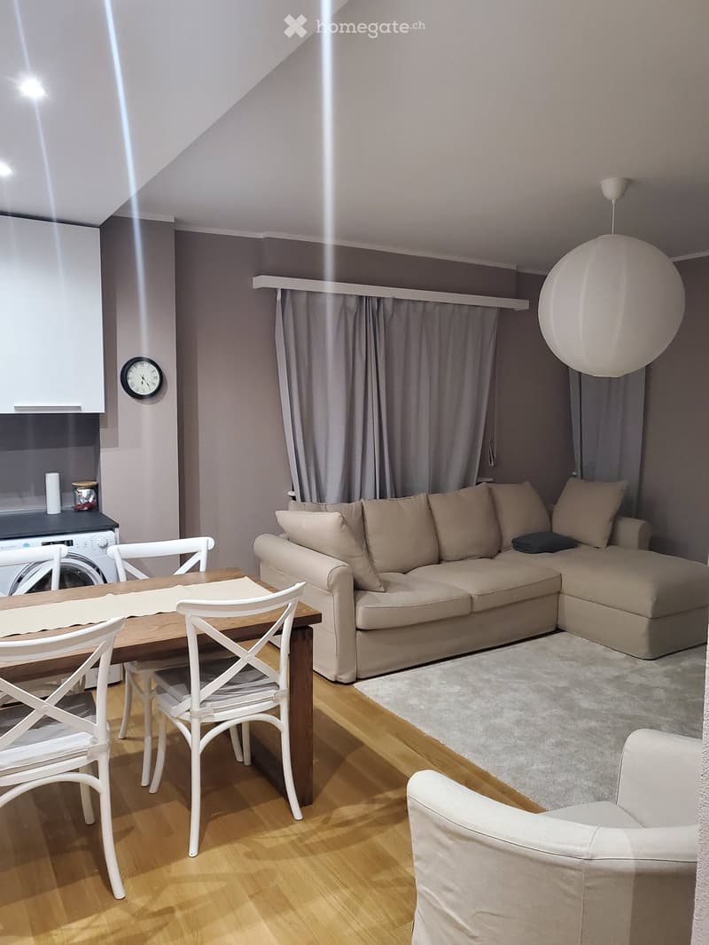 A modern, well-lit living room with a beige sectional sofa, a wooden dining table with white chairs, a white armchair, a washing machine, and a large round pendant light.