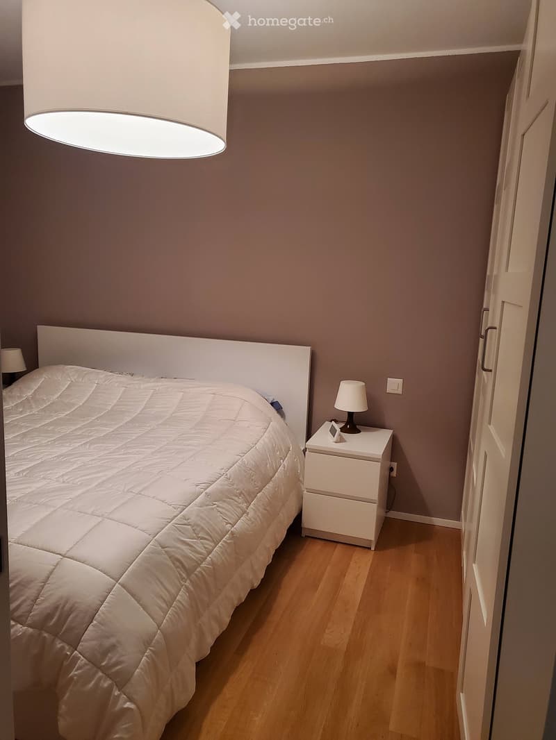 A modern bedroom with a large bed, white headboard, nightstand, and lamp. The room features wooden flooring and neutral-colored walls.