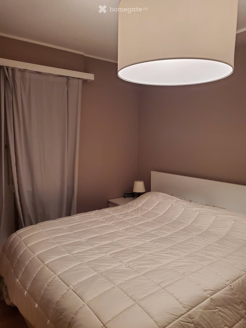 A neatly made bed with a white duvet and a matching headboard, positioned against a gray wall. A nightstand with a lamp sits beside the bed, and a curtain is visible on the left side.