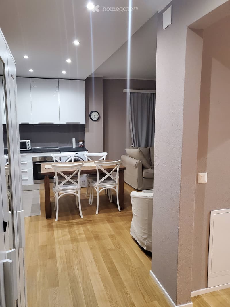 Open-plan living and dining area with light wooden flooring, white cabinets, a sofa, a dining table with chairs, and a microwave in the kitchen area.