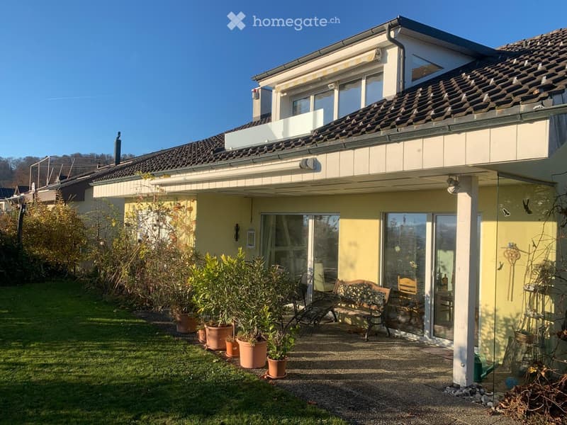 Two-story house with a tiled roof, large windows, and a balcony on the upper level. The front yard features a well-maintained lawn, several potted plants, and outdoor seating including a bench and chairs.