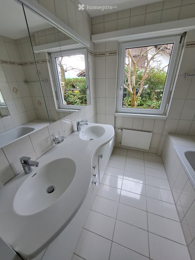 Modern bathroom with white tiles, a large sink with two faucets, a bathtub, and a large mirror. Natural light streams in through two windows showing a garden view.
