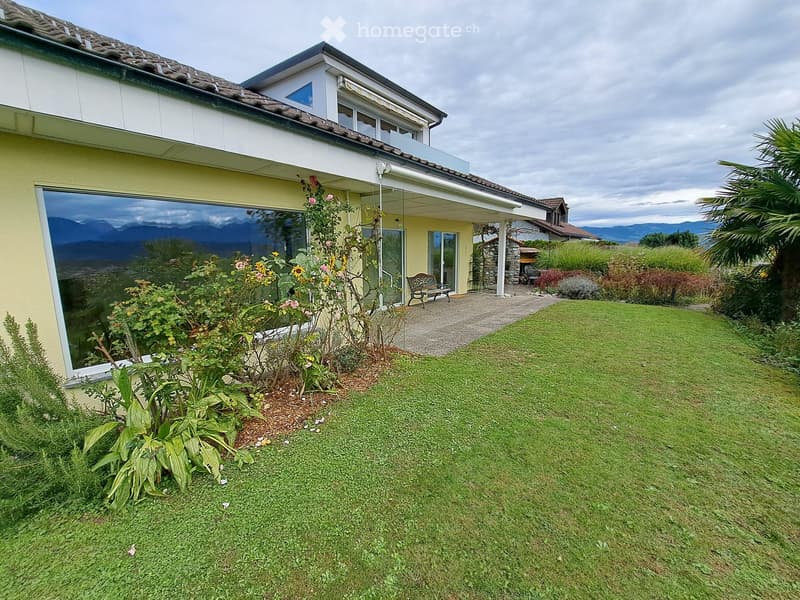 Well-maintained garden with a lawn, flower beds, and a bench near the house entrance. The house has large windows and a tiled roof, with a scenic mountain view in the background.