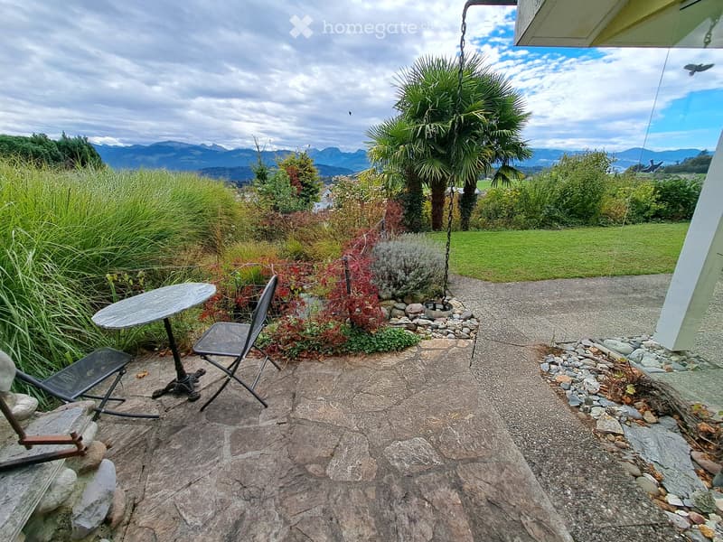 Outdoor seating area with a round table and two chairs on a stone patio, surrounded by lush greenery and a scenic mountain view.