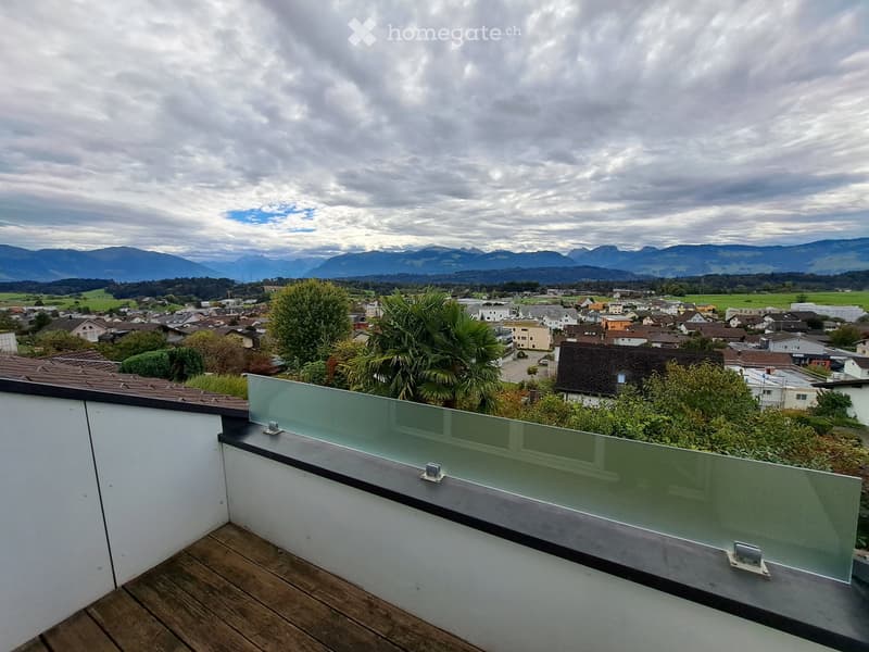 A modern rooftop terrace with wooden decking, glass railing, and scenic views of a town and distant mountains.