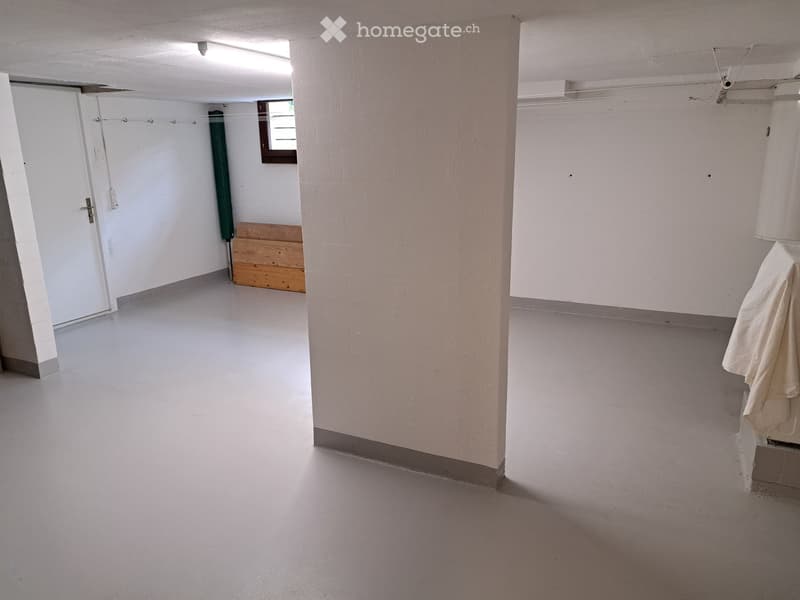 Empty room with white walls, grey floor, and a small wooden bench in the corner. A white door is visible on the left side, and a window is seen in the background.