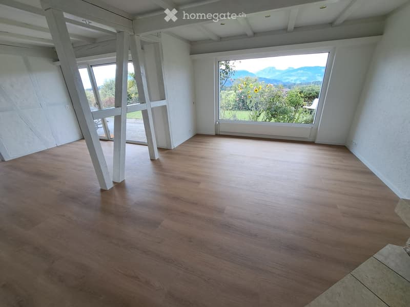 Empty room with wooden flooring, white walls, large windows, and visible beams in the ceiling.