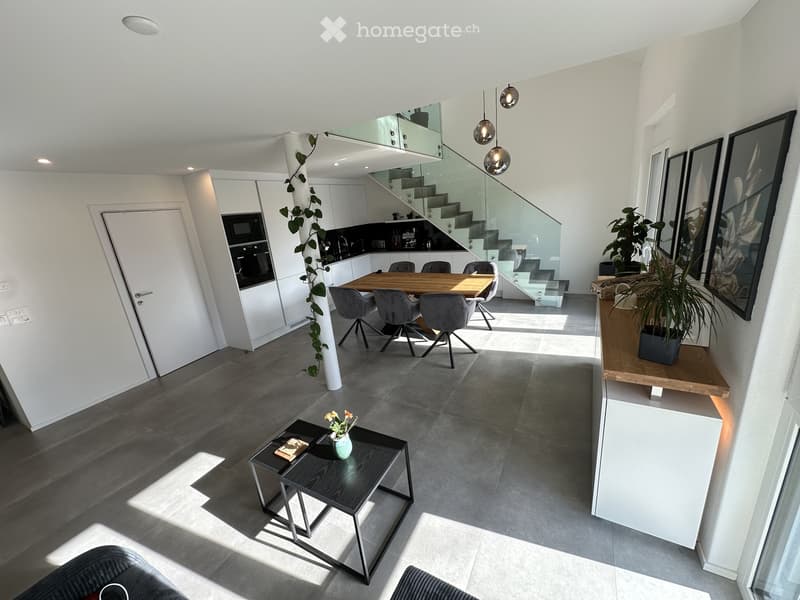 Modern open-plan living and dining area with grey sofa, coffee table, dining table with chairs, pendant lights, and a staircase leading to an upper level.
