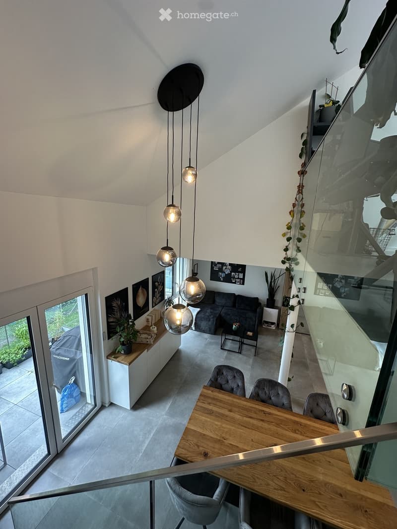 Modern living room with a grey sofa, coffee table, wooden dining table, pendant lights, and a glass staircase leading to an upper level.