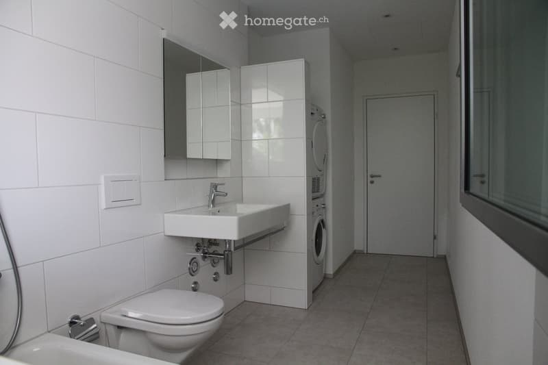 Modern bathroom with white tiles, a sink, toilet, mirror, and a washing machine in the corner.