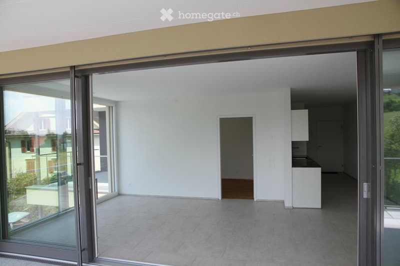 Empty room with large sliding glass doors, tiled floor, white walls, and a small kitchen area visible in the background.