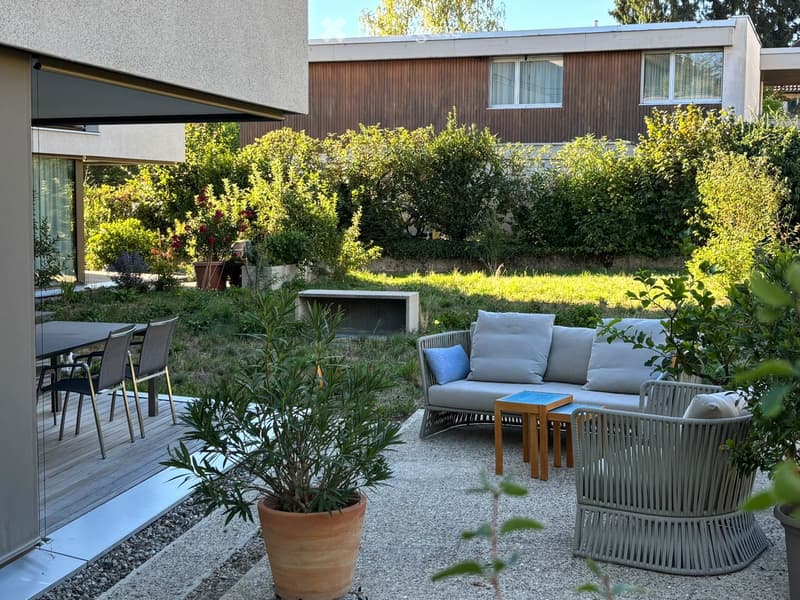 Modern outdoor seating area with a grey sofa, wooden coffee table, wicker chairs, and potted plants, surrounded by greenery and a modern building.