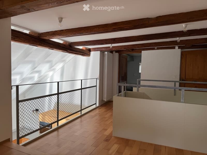 An empty attic space with wooden beams, exposed wooden ceiling, and wooden flooring. Features include a metal railing and a skylight window allowing natural light to enter.