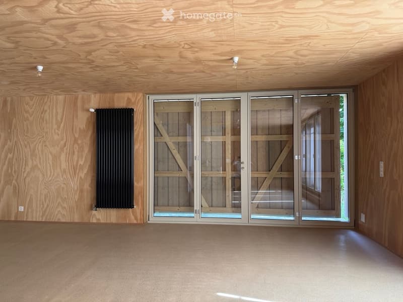 Empty room with wooden walls and ceiling, large glass sliding doors, and a black radiator on one wall.