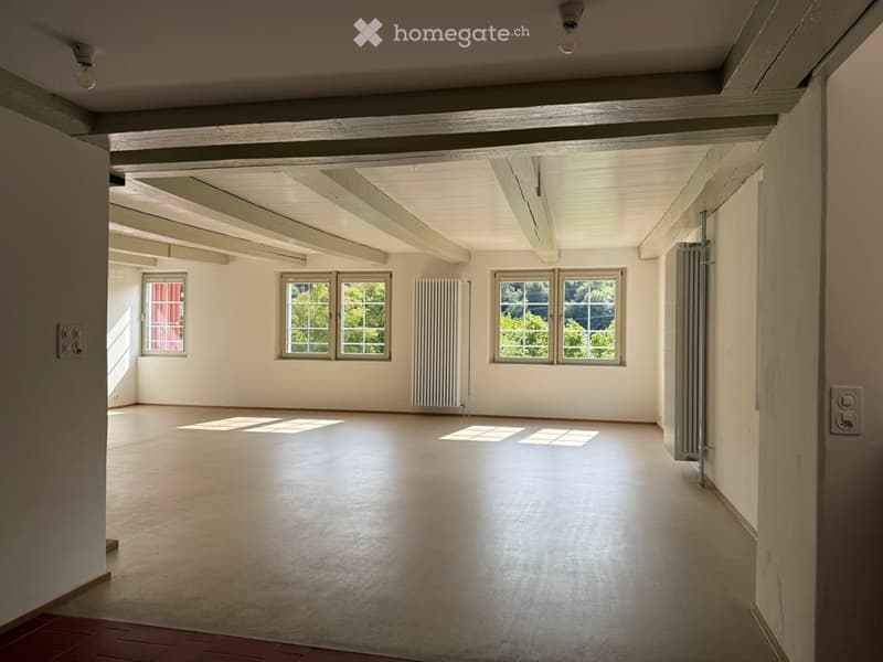 Empty room with white walls, wooden ceiling beams, large windows, and a smooth concrete floor.