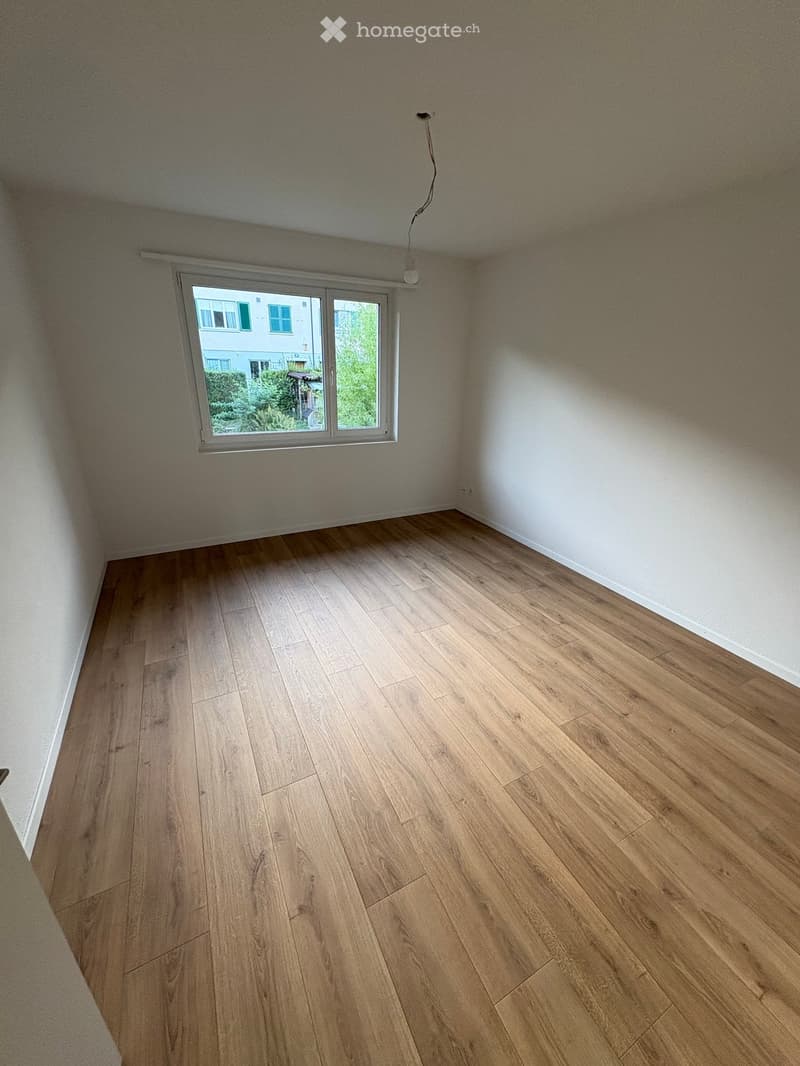 Empty room with light wooden flooring and white walls, featuring a large window with a view of greenery and neighboring buildings.
