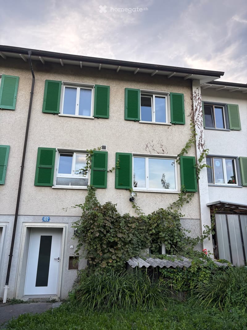 Two-story house with light-colored facade, green shutters, multiple windows, and climbing vines on the wall. The entrance has a white door with a number sign above it.
