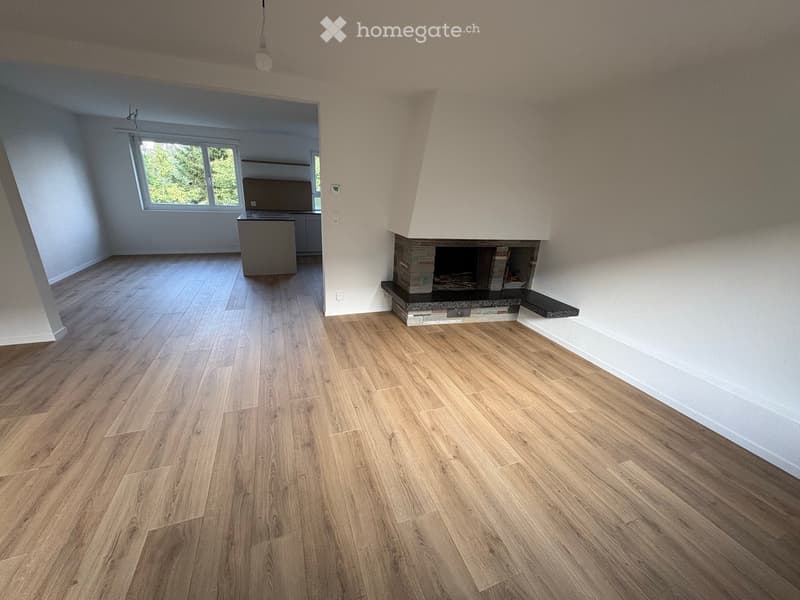 Empty room with wooden flooring, white walls, and a fireplace. There is a window with a view of greenery and a small kitchen area visible in the background.