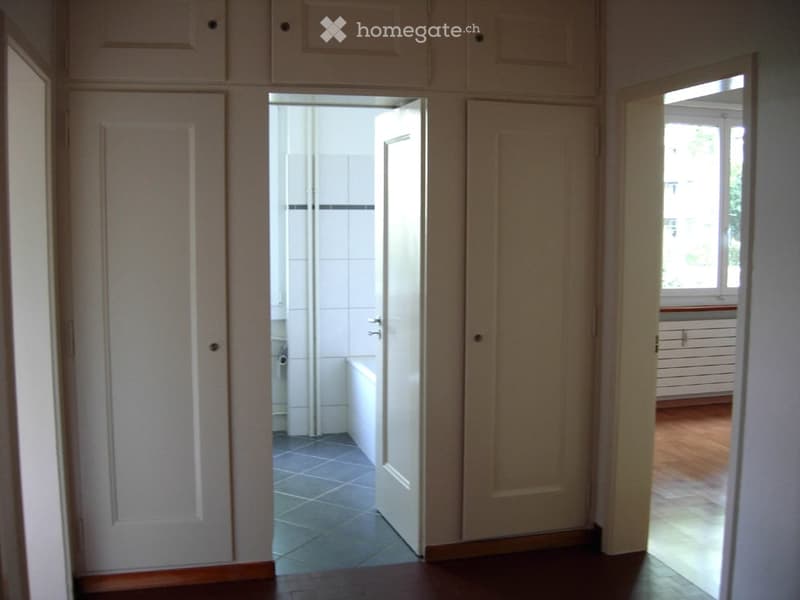 A narrow hallway with wooden flooring, white walls, and several closed doors leading to different rooms. The hallway has a tiled floor in the adjacent bathroom area visible through an open door.