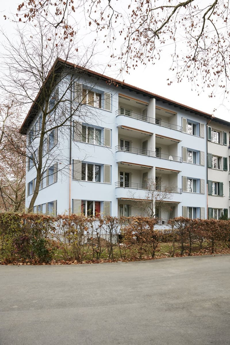 A modern, light blue multi-story building with multiple balconies and large windows, surrounded by bare trees and shrubs.