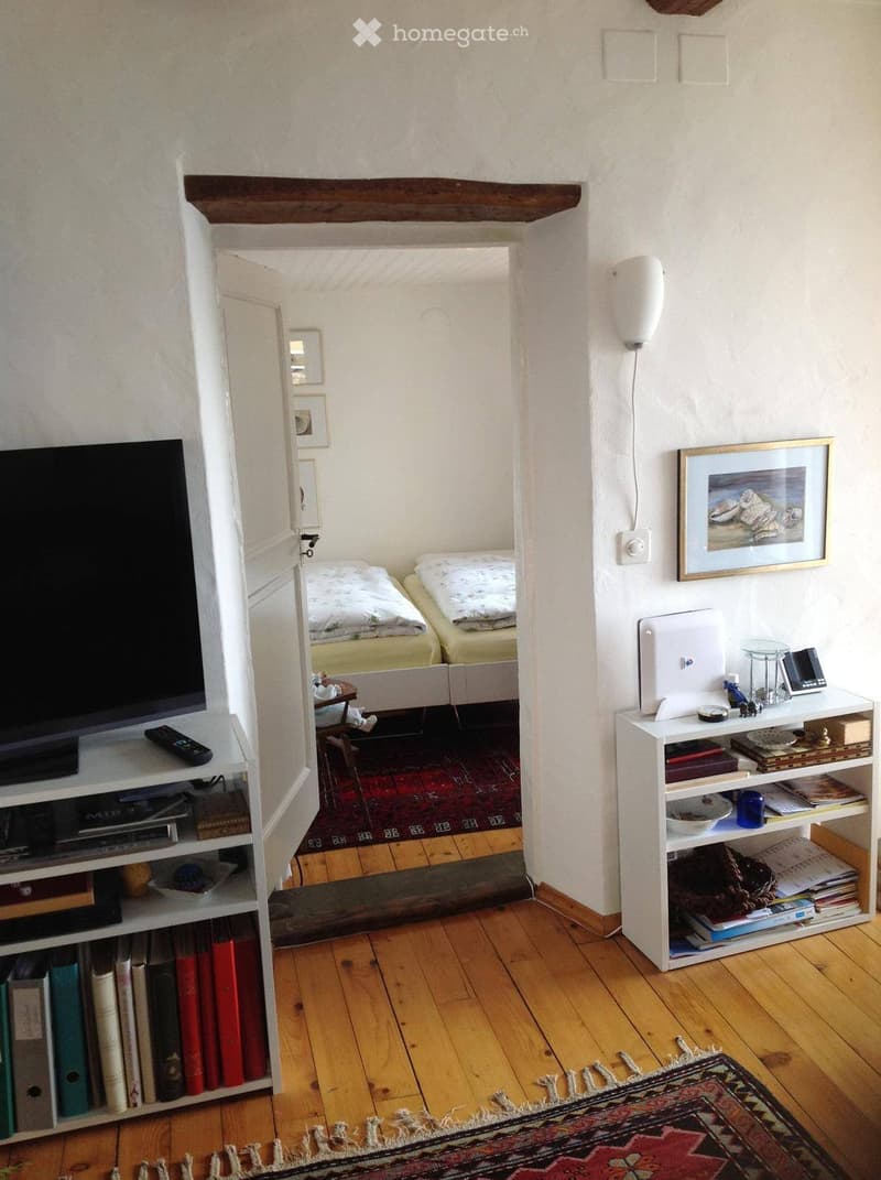 A cozy living space with a TV on a shelf, a small table with a bed visible in the background, and shelves filled with books and decorative items.