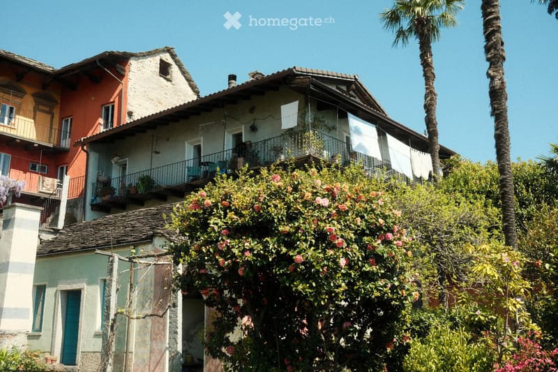 A multi-story residential building with a tiled roof, balconies, and surrounding greenery, including palm trees and flowering bushes.