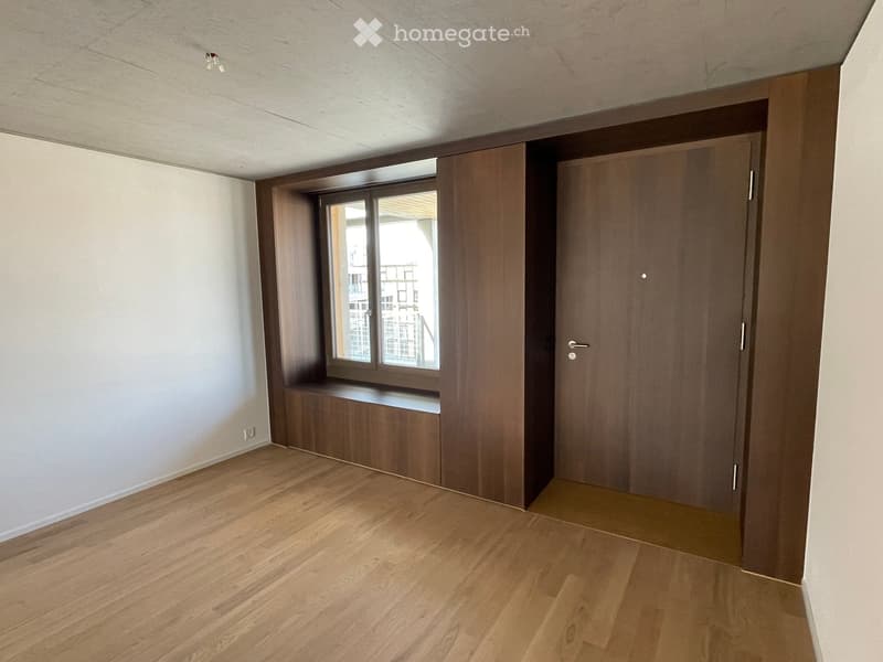Empty room with wooden flooring, white walls, a large window with a built-in bench, and a closed wooden door.