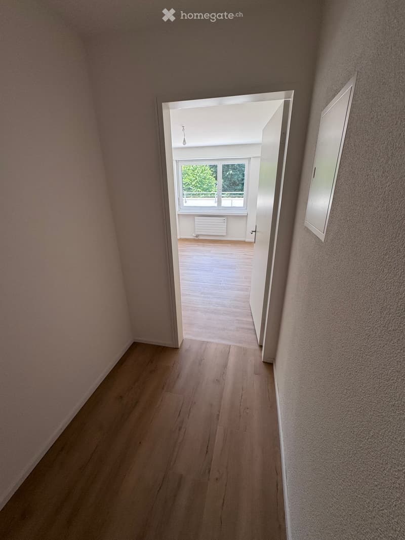 Empty hallway with wooden flooring, light-colored walls, and an open doorway leading to another room with a window and radiator.